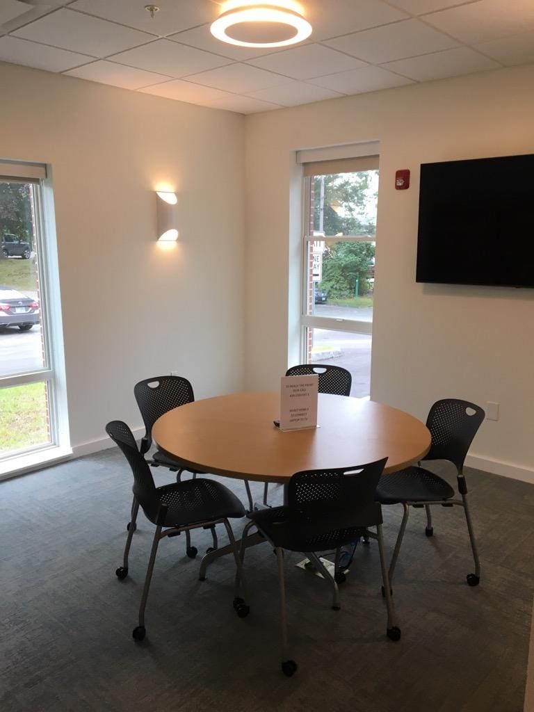 A small meeting room with a round table, five black chairs, large windows, a wall-mounted TV, and a sign on the table. Natural light fills the room.