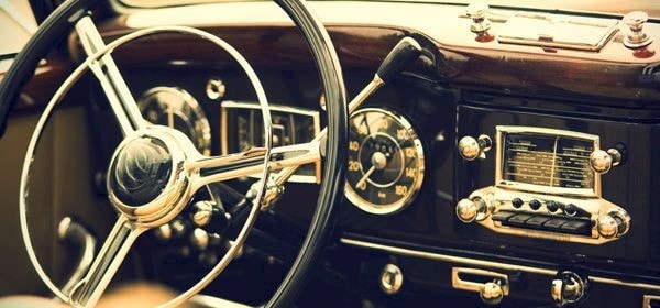 Close-up of a vintage car dashboard featuring a large steering wheel, analog speedometer, radio with buttons and dials, and a gear shift lever. The interior has a classic, polished design.