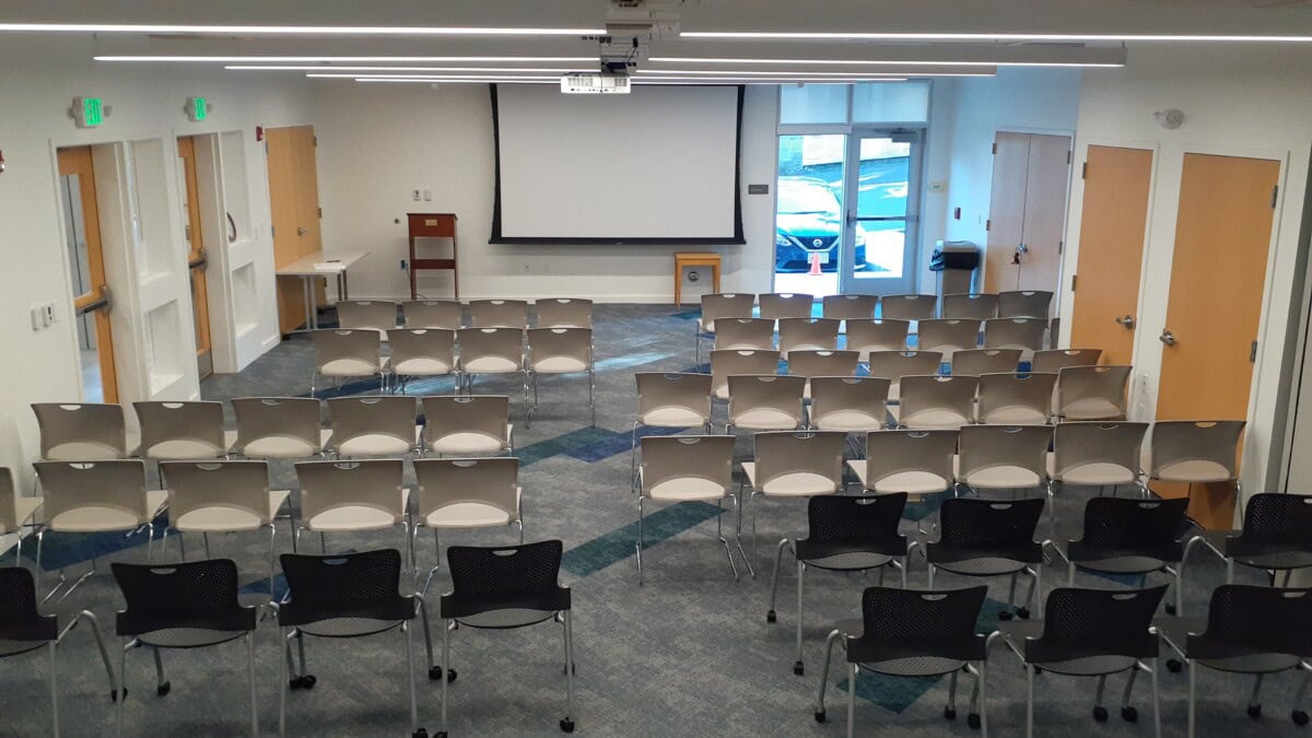 A conference room with rows of empty chairs facing a large projection screen, doors on the left and right walls, windows at the back, and a table near the screen at the front.
