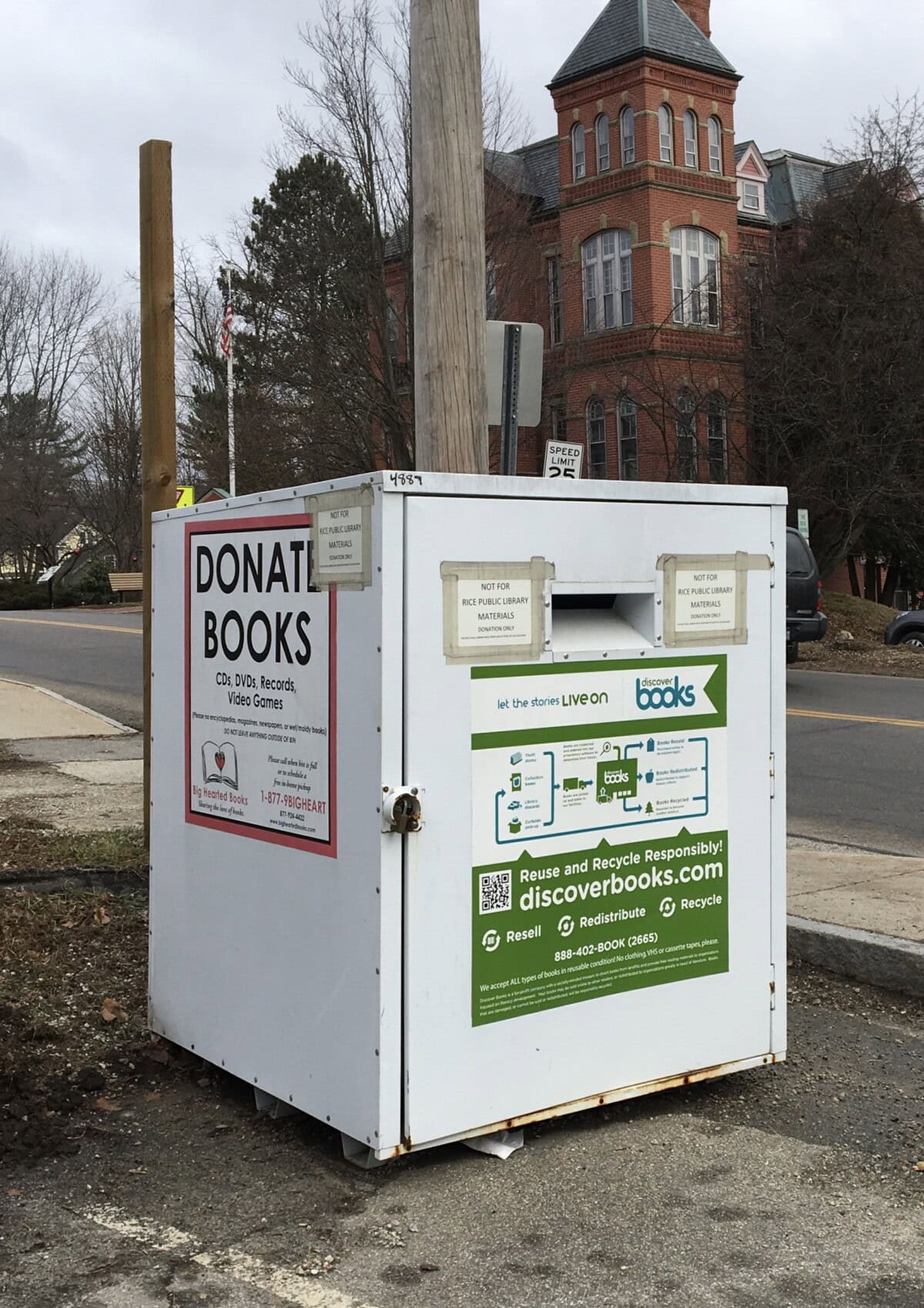 A white outdoor donation bin for books stands on a sidewalk near a street, with signs for donating CDs, DVDs, records, and video games. In the background, there are leafless trees and a large brick building.
