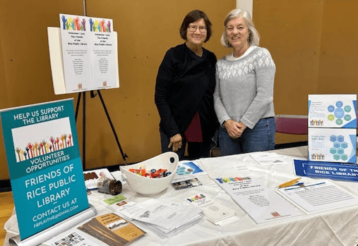 Two women stand behind a table with library flyers, brochures, and a candy bowl at an event. Signs on the table promote volunteer opportunities at the Friends of Rice Public Library.