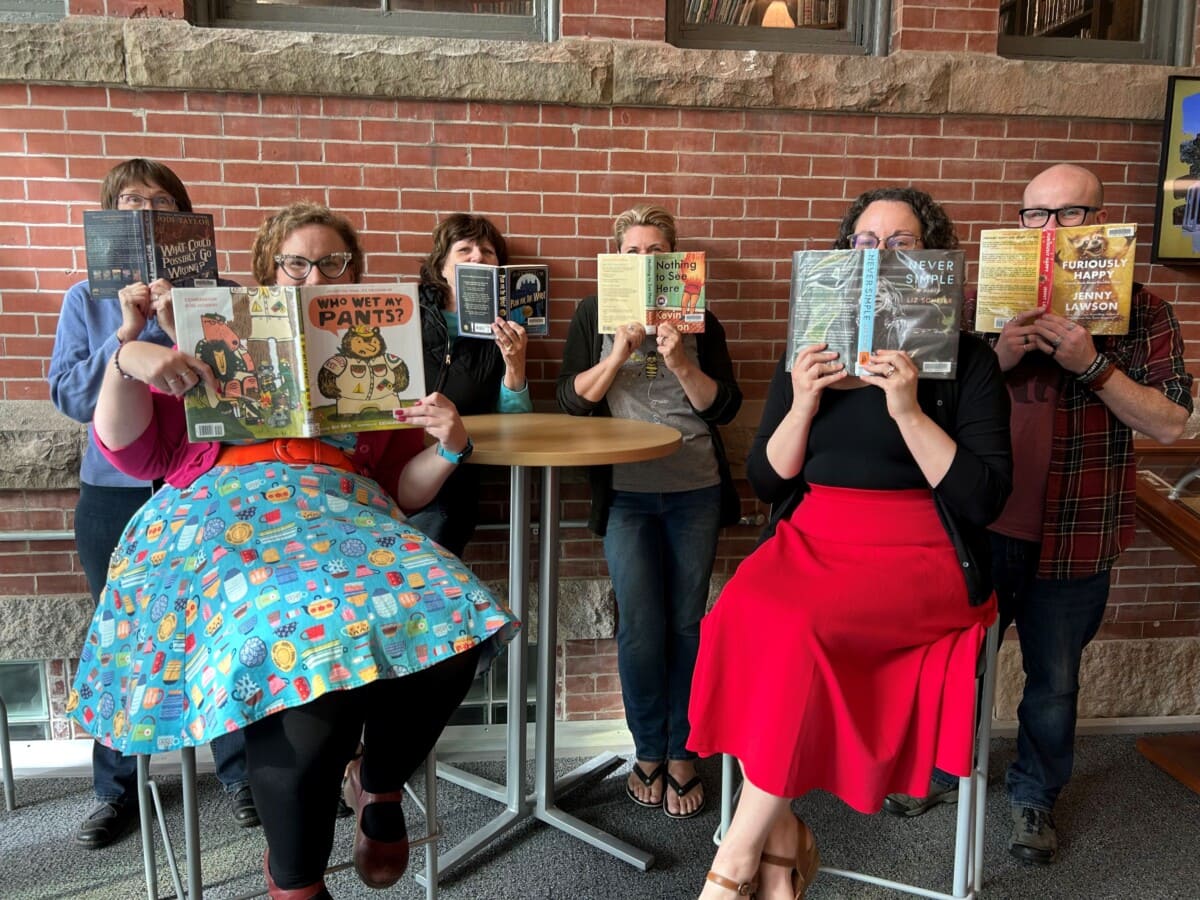 Six adults sitting and standing around a round table in front of a brick wall, all holding books or graphic novels that cover their faces and appear to be reading or posing for a playful group photo.
