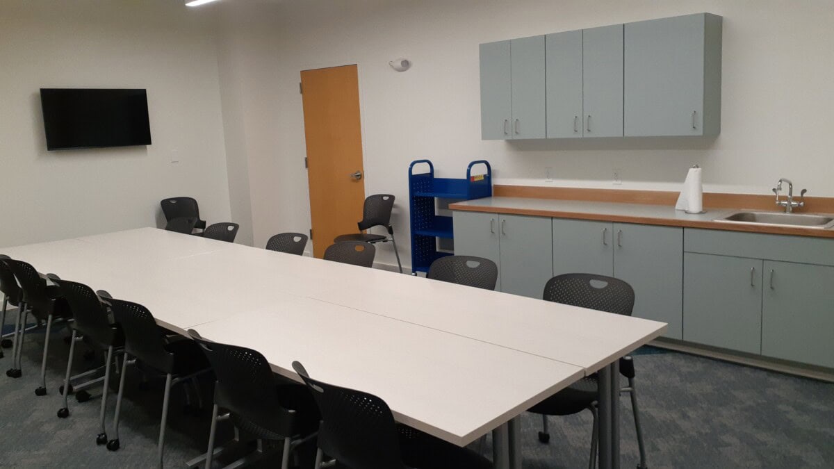 A conference room with a long table surrounded by chairs, a wall-mounted TV, a rolling cart, light gray cabinets, a counter with a sink, and a paper towel roll on the counter.