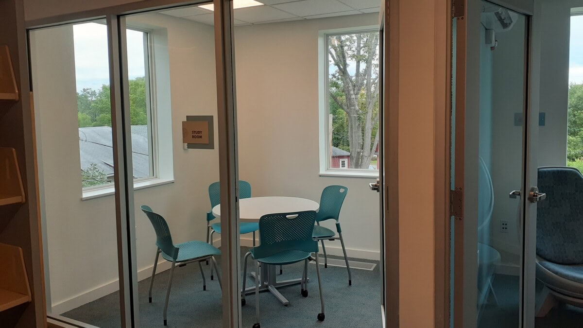 A small study room with a round table and four teal chairs, enclosed by glass walls. There are two large windows letting in natural light, and trees are visible outside. A sign on the wall reads STUDY ROOM.
