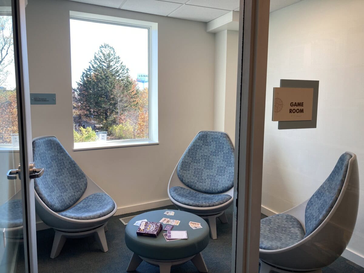 A small room with four modern blue chairs arranged in a circle, a round table in the center with playing cards and games, a window with trees outside, and a sign on the door that reads Game Room.